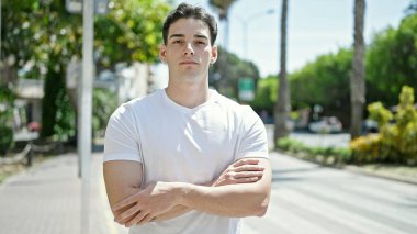 Ciddi bir ifade ve kollarını kavuşturarak parkta dikilen İspanyol genç bir adam.