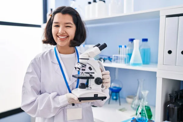 Joven Mujer Hispana Trabajando Laboratorio Científico Sosteniendo Microscopio Guiñando Ojo —  Fotos de Stock
