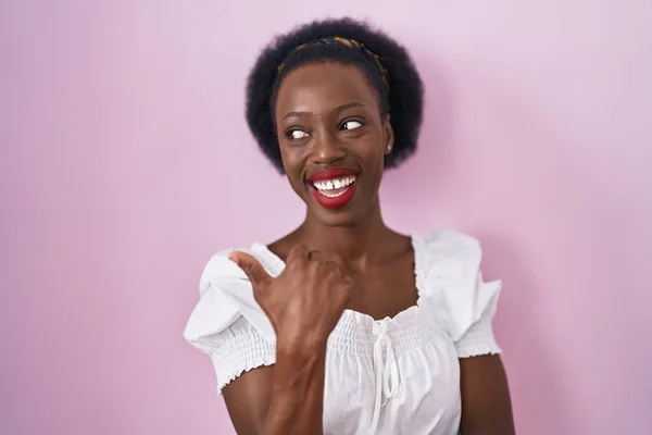 头发卷曲的非洲妇女站在粉红的背景上 笑容满面 手指头指向侧面 — 图库照片
