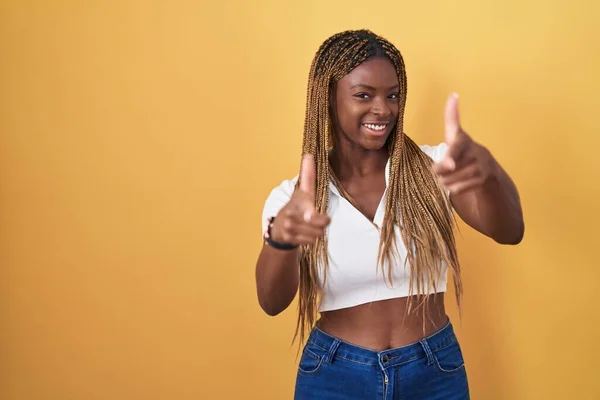 非洲裔美国妇女 辫子式的头发 站在黄色的背景上 手指指向镜头 笑容可亲 充沛的精力和活力 — 图库照片