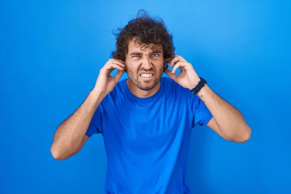 青い背景の上に立つヒスパニック系の若者は 大きな音楽のノイズのためにいらいらする表情で指で耳を覆います 聴覚障害の概念 — ストック写真