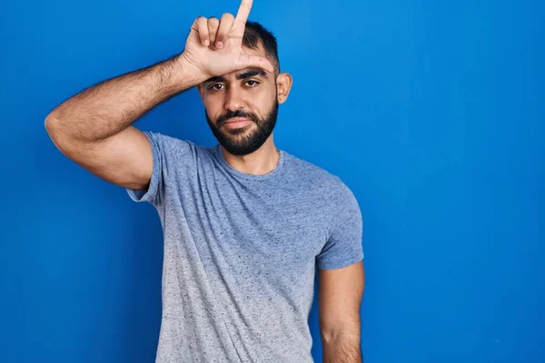 中东男人留着胡子 站在蓝色的背景上取笑额头上的人做失败者的手势 嘲笑和侮辱 — 图库照片
