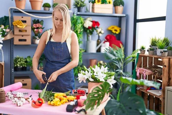 Young blonde woman florist cutting stem at florist store