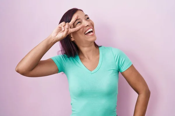 ピンクの背景の上に立つ若いヒスパニック系の女性は 顔の上に指で平和のシンボルを行います 勝利を示す明るい笑顔 — ストック写真