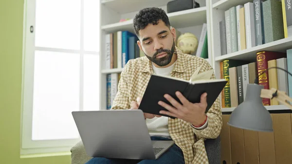 Young hispanic man student using laptop reading book at library university