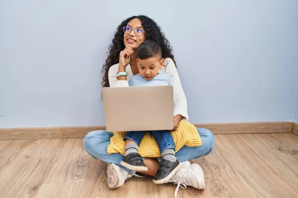 若いヒスパニック系の母親と子供のコンピュータのラップトップの床に手であごの質問については 集中的な表現を考えて座って使用しています 笑顔で思いやりのある顔 疑わしい概念 — ストック写真