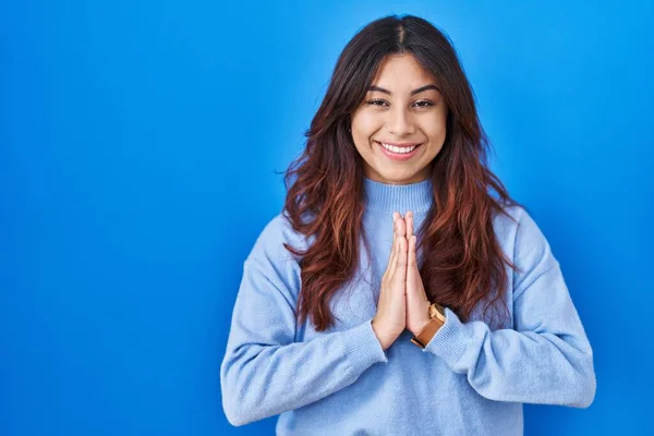 西班牙裔年轻女子站在蓝色的背景上手拉手祈祷 祈求宽恕 充满自信地微笑着 — 图库照片