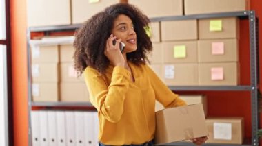 Afrika kökenli Amerikalı kadın ekommerce işçisi elinde paketle ofisteki akıllı telefondan konuşuyor.