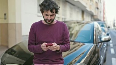 Young hispanic man using smartphone leaning on car at street