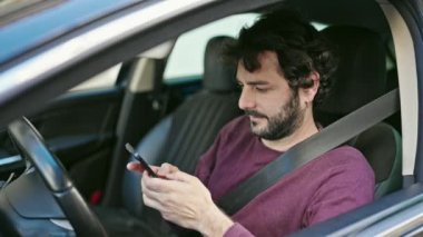 Young hispanic man using smartphone sitting on car at street