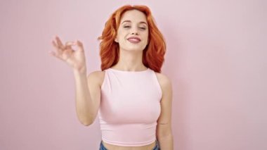 Genç kızıl saçlı kadın pembe arka planda güzel bir jestle gülümsüyor.