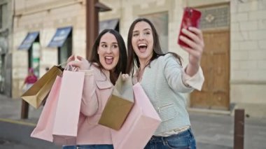 Alışverişe giden iki kadın ellerinde çantalarla sokakta akıllı telefondan selfie çekiyorlar.