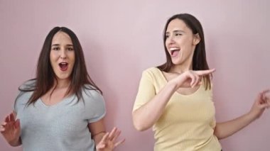 İki kadın gülümsüyor pembe arka planda dans ediyorlar.