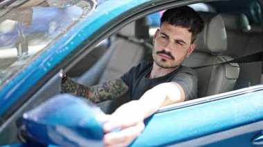 Latin kökenli genç adam dikiz aynasına dokunuyor. Sokakta arabada oturuyor.