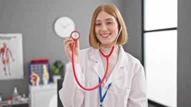 Klinikte steteskop tutan genç sarışın doktor gülümsüyor.