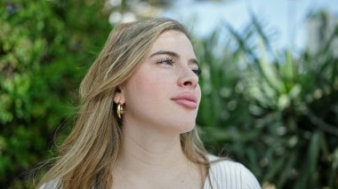 Genç sarışın kadın parkta rahat bir ifadeyle yan tarafa bakıyor.