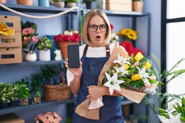 Çiçekçide çalışan beyaz bir kadın şok olmuş bir suratla akıllı telefon ekranı gösteriyor. Şüpheci ve alaycı görünüyor. 