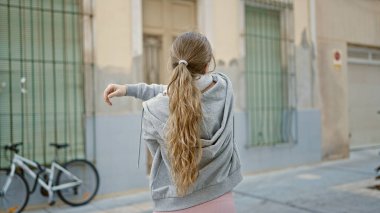 Spor kıyafetleri giyen genç sarışın kadın caddede kollarını esnetiyor.