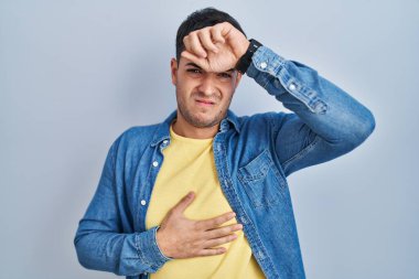 Young hispanic man standing over blue background touching forehead for illness and fever, flu and cold, virus sick 
