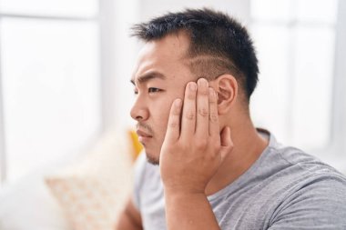 Young chinese man suffering for ear pain sitting on bed at bedroom