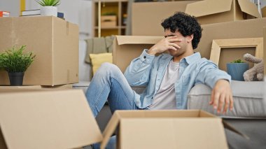 Genç latin adam yeni evinde yerde stresli oturuyor.