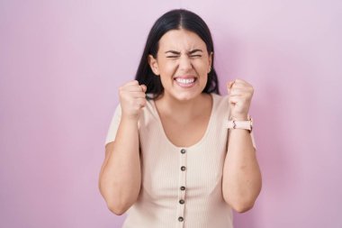 Pembe arka planda duran genç İspanyol kadın başarı için heyecanlı kolları havada ve gözleri kapalı zaferi kutlarken gülümsüyor. Kazanan konsept. 