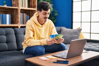 Young caucasian man using laptop counting swiss franc banknotes at home