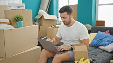 Young hispanic man using laptop sitting on sofa at new home