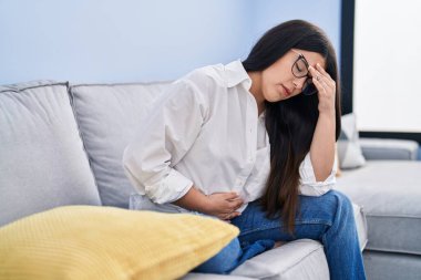 Young hispanic woman sitting on sofa suffering for stomach pain at home