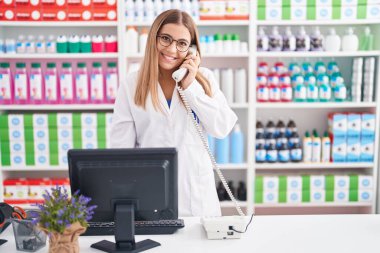 Young beautiful hispanic woman pharmacist talking on telephone using computer at pharmacy