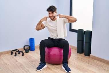 Yoga odasında pilatese oturmuş sakallı, neşeli gülümseyen ve parmaklarıyla ve ağzıyla işaret eden İspanyol adam. diş sağlığı kavramı. 