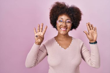 Pembe arka planda duran genç Afro-Amerikalı kadın dokuz numarayı gösterip işaret ederken kendinden emin ve mutlu bir şekilde gülümsüyor.. 