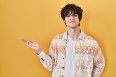 Genç adam rahat bir yaz gömleği giyer neşeli bir şekilde sunar ve avuç içiyle kameraya bakar.. 