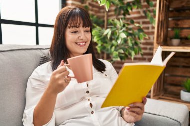 Young beautiful hispanic woman reading book and drinking coffee sitting on sofa at home