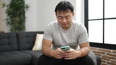 Young chinese man using smartphone sitting on sofa at home