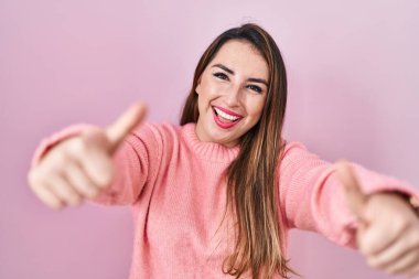Young hispanic woman standing over pink background approving doing positive gesture with hand, thumbs up smiling and happy for success. winner gesture. 