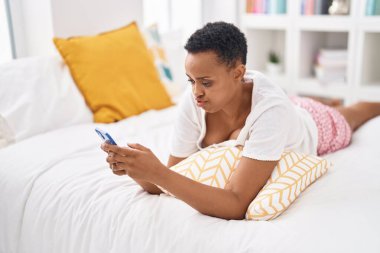 Afrika kökenli Amerikalı kadın yatak odasında endişeli bir ifadeyle akıllı telefon kullanıyor.