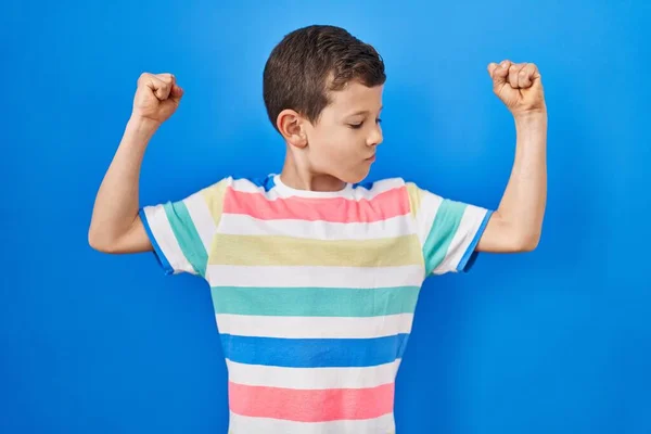 青い背景に立っている若い白人の子供は腕の筋肉が誇らしげに笑っているのを示しています フィットネスのコンセプト — ストック写真