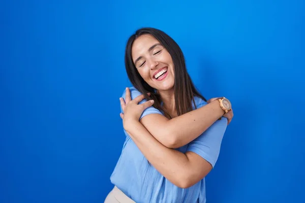 青の背景に立っている若いブルネットの女性は自分自身を幸せと肯定的に抱き 自信を持って笑っています 自己愛と自己ケア — ストック写真