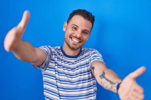 青い背景の上に立っている若いヒスパニック系の男が抱擁のために開いて腕で笑みを浮かべてカメラを見て 幸せを受け入れる陽気な表情 — ストック写真