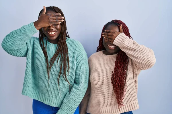 青い背景の上に立つ2人のアフリカ人女性が笑顔で 驚きのために目を覆う顔で笑っています 視覚障害の概念 — ストック写真