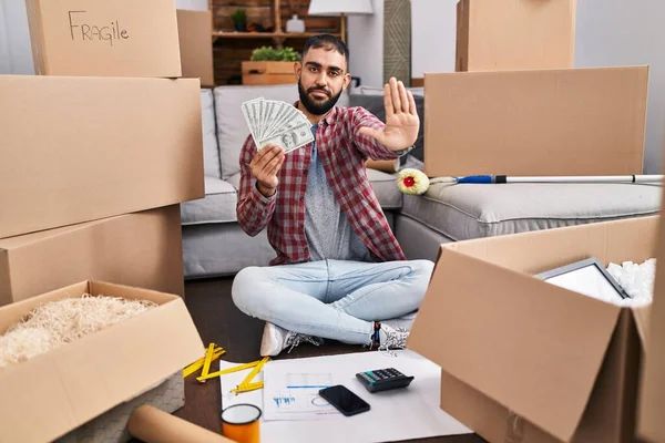 Middle east man with beard sitting on the floor at new home holding money with open hand doing stop sign with serious and confident expression, defense gesture