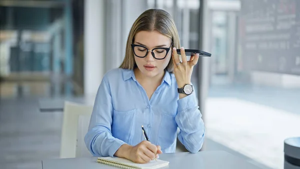 Young blonde woman business worker listening voice message by smartphone taking notes at office