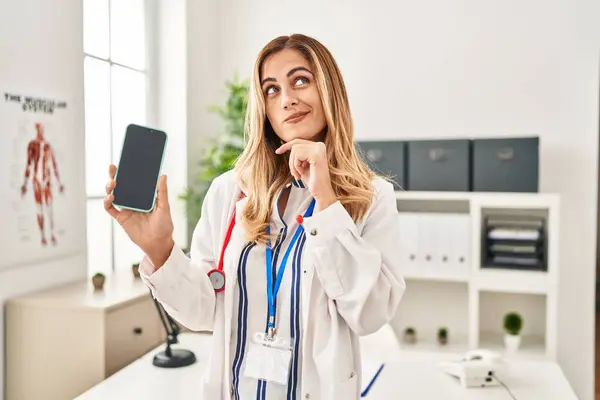 Young Blonde Doctor Woman Working Clinic Showing Smartphone Screen Serious — Stock Photo, Image