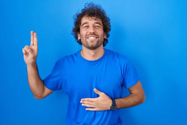 西班牙裔年轻人站在蓝色的背景上微笑着 手托着胸膛和手指发誓 宣誓效忠 — 图库照片