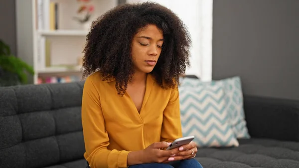アフリカ系アメリカ人の女性が自宅のソファに座ってスマートフォンを使って — ストック写真