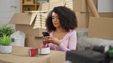 Afro-Amerikalı kadın akıllı telefon kullanıyor. Yeni evinde koltukta oturmuş kutlama yapıyor.