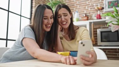 Kahve içen iki kadın yemek odasında akıllı telefondan video izliyorlar.