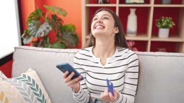 Genç, İspanyol bir kadın akıllı telefon ve kredi kartıyla alışveriş yapıyor. Evdeki koltukta oturuyor.