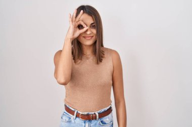Young hispanic woman standing over white background doing ok gesture with hand smiling, eye looking through fingers with happy face. 